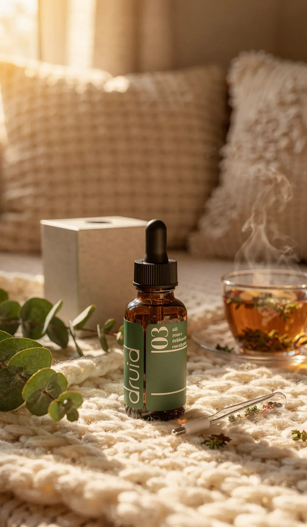 1 flacon brun avec compte-gours vert et étiquette Potion 03 Défenses Immunitaires Druid, pipette et tasse de thé fumante sur un tapis doux, ambiance cocooning.