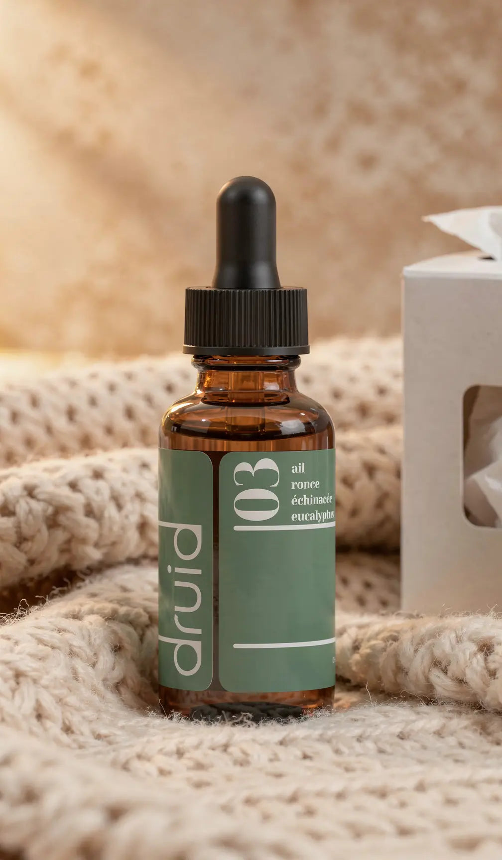 Goutte-à-goutte brun dans flacon en verre avec étiquette vert pastel portant “druid” et ingrédients ail, rome, échinacée, eucalyptus, posé sur une laine crème.