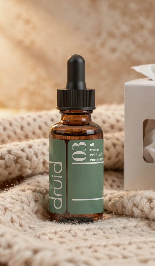 Goutte-à-goutte brun dans flacon en verre avec étiquette vert pastel portant “druid” et ingrédients ail, rome, échinacée, eucalyptus, posé sur une laine crème.