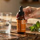 Goutte d’élixir naturelle dans un flacon brun, tenus au doigt, à côté d’un verre d’eau et de feuilles de menthe sur une table en bois.